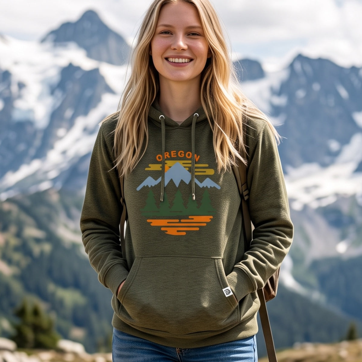 A smiling woman outdoors in front of snowy mountains wears an olive green pullover hoodie with a colorful 'OREGON' mountain-and-lake graphic on the chest.