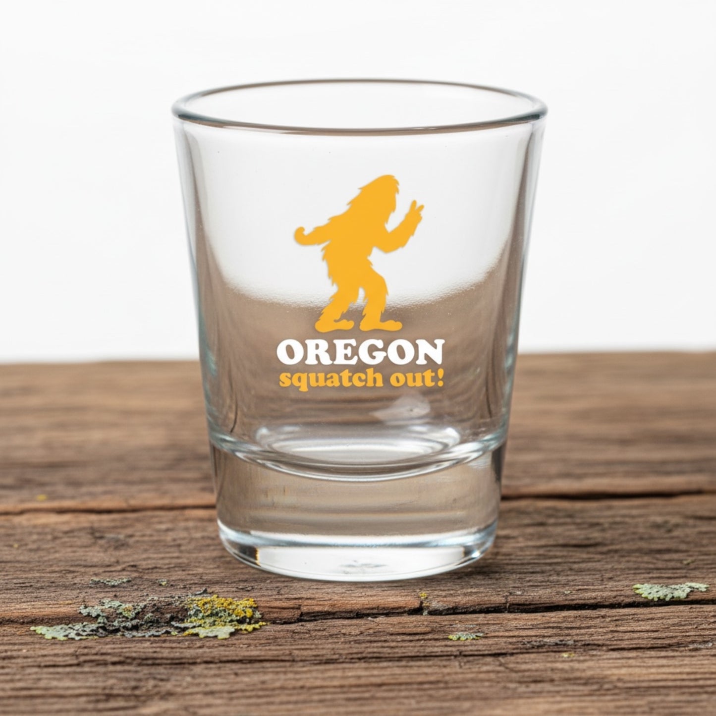 A clear glass shot tumbler with a yellow and white graphic of Bigfoot and the text 'OREGON squatch out!' sitting on a rustic wooden surface.