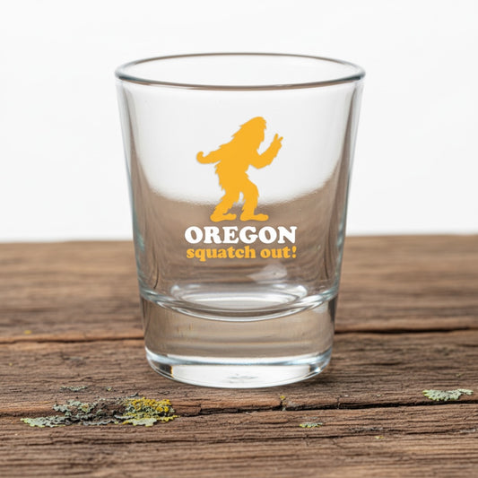 A clear glass shot tumbler with a yellow and white graphic of Bigfoot and the text 'OREGON squatch out!' sitting on a rustic wooden surface.