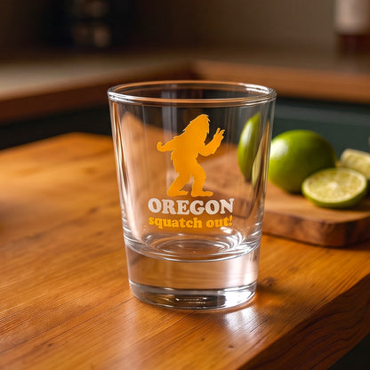 A clear glass tumbler with a yellow graphic of Bigfoot and the text 'OREGON squatch out!' sits on a wooden table, with limes blurred in the background.