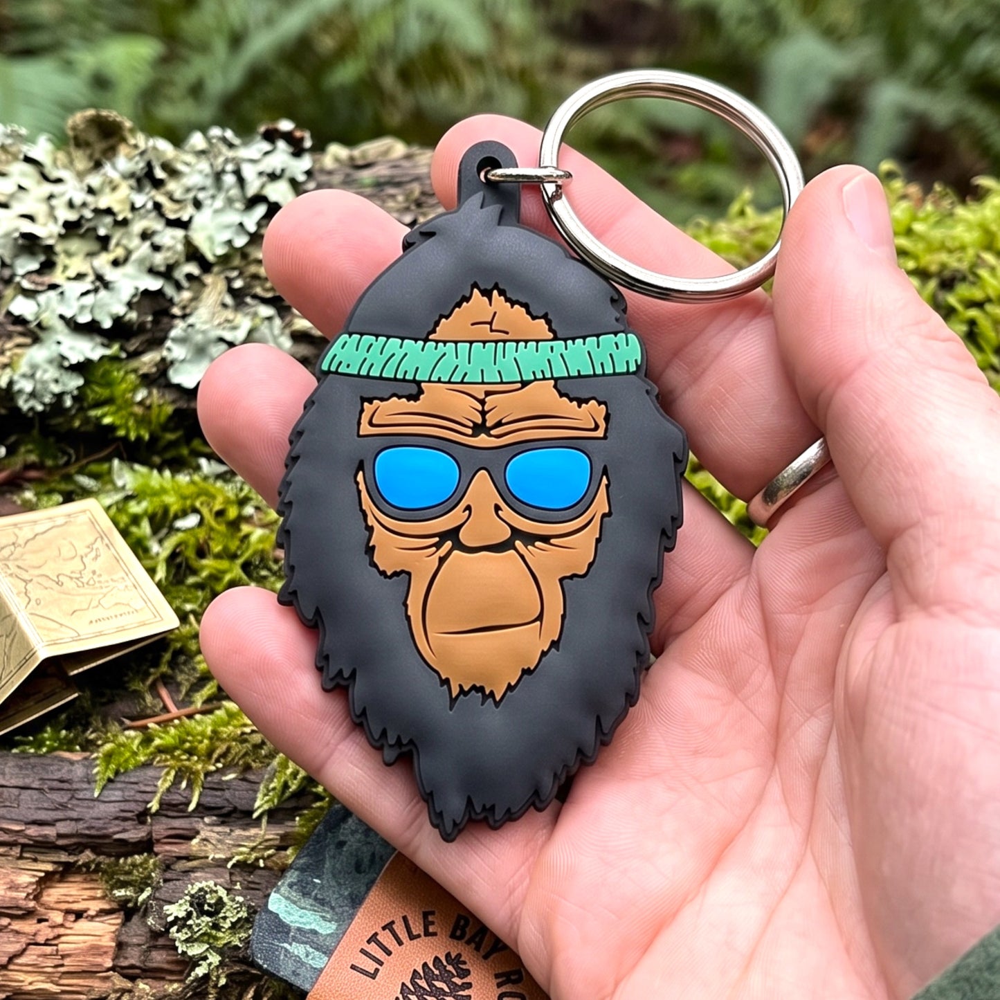A hand holds a rubber keychain shaped like a gorilla head with a black beard, brown face, blue sunglasses, and a green headband, attached to a silver metal key ring. The background is an outdoor mossy forest setting.