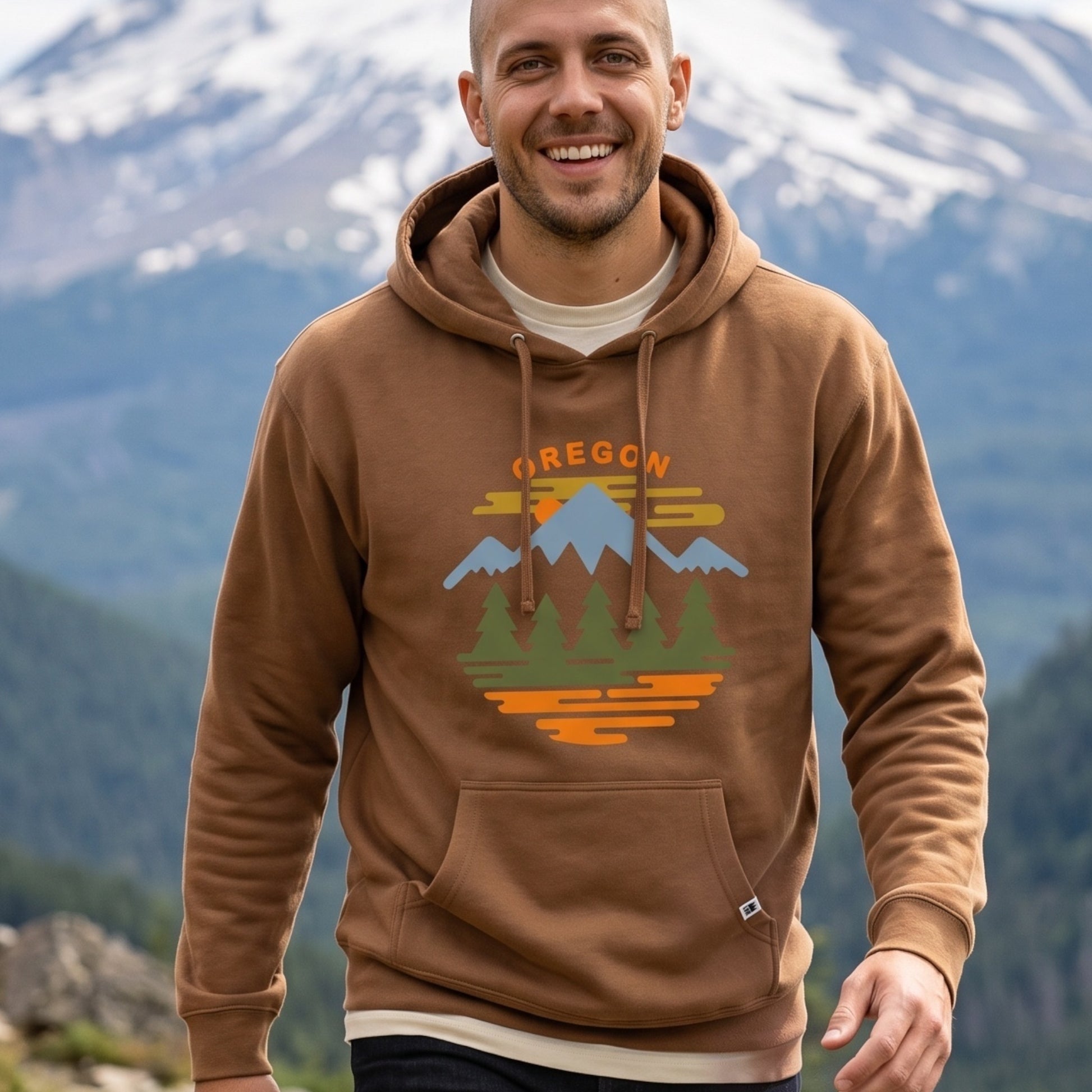 A smiling man wearing a brown pullover hoodie with a colorful 'OREGON' mountain graphic on the chest, set against a blurred background of snow-capped mountains and forests.