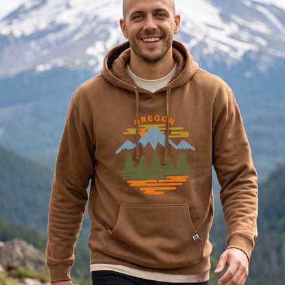 A smiling man wearing a brown pullover hoodie with a colorful 'OREGON' mountain graphic on the chest, set against a blurred background of snow-capped mountains and forests.