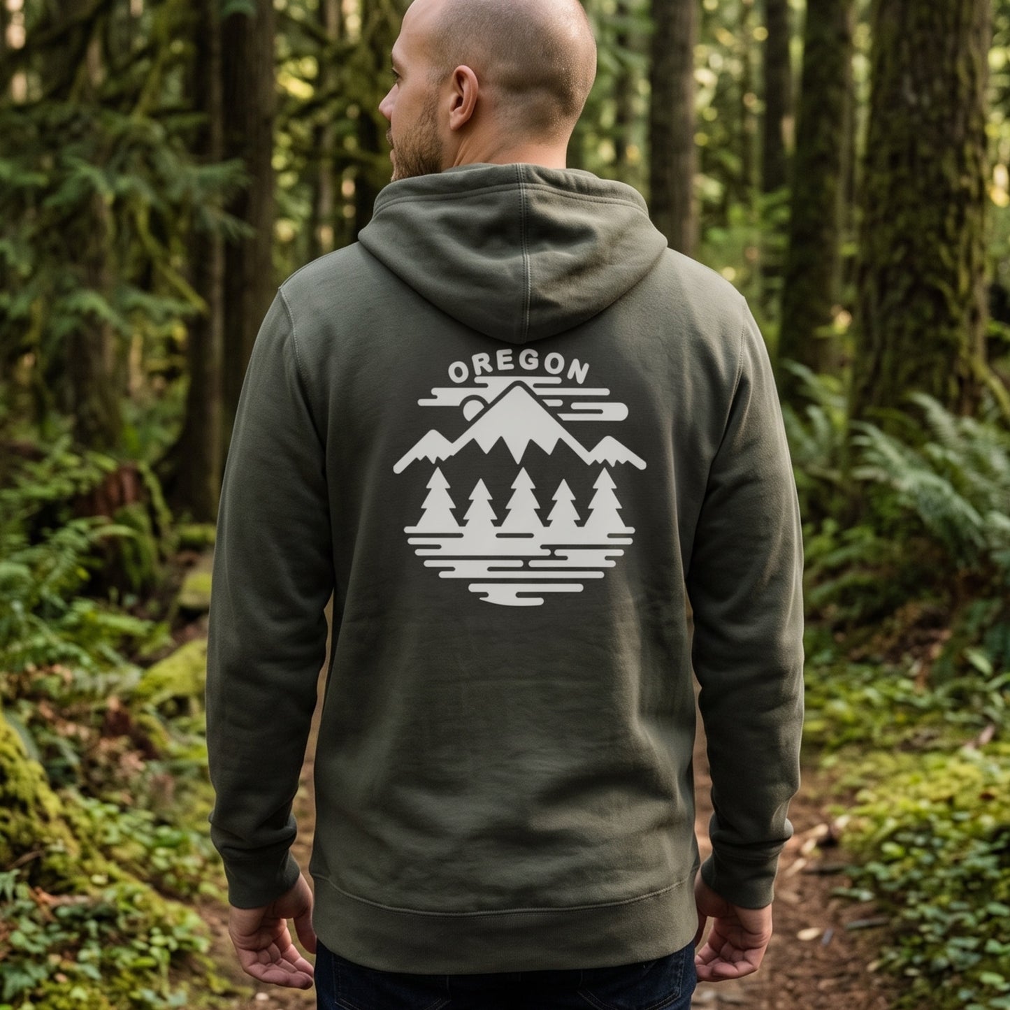 A man viewed from behind wearing an olive green hoodie with a white 'OREGON' mountain and tree graphic on the back, standing in a forest.