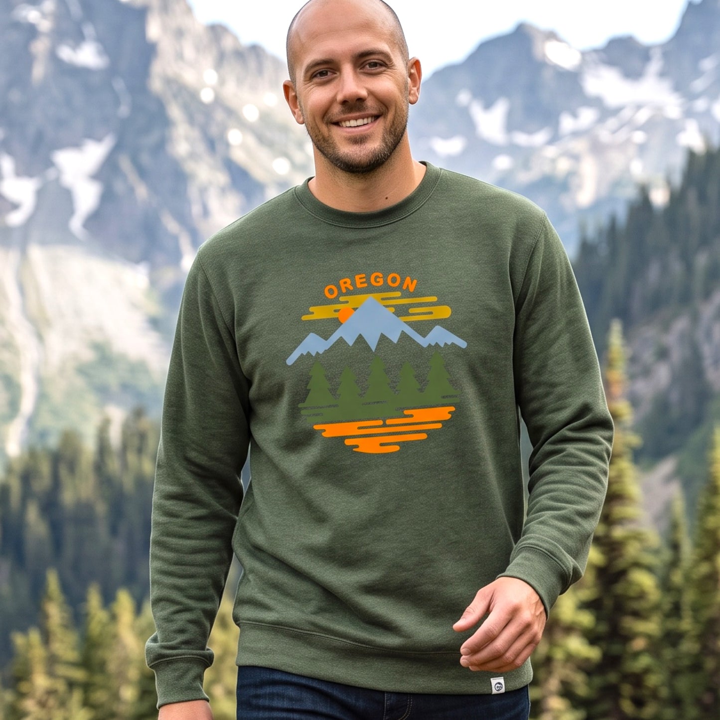 A smiling man wearing an olive green crewneck sweatshirt with a colorful 'OREGON' mountain landscape graphic, set against a background of pine trees and snow-capped mountains.