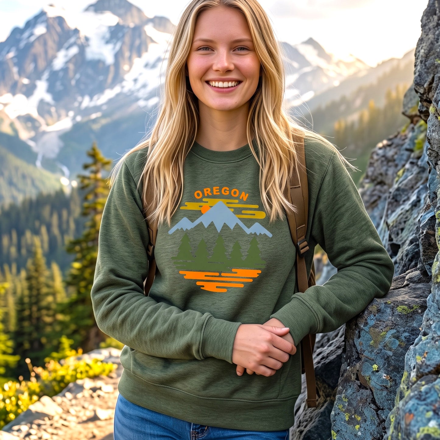 A smiling blonde woman wearing an olive green crewneck sweatshirt with a colorful 'OREGON' mountain and tree graphic, standing outdoors against a mountain landscape background.