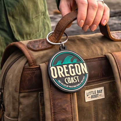 A close-up of a round black and teal 'Oregon Coast' keychain hanging from a brown leather handle on a tan canvas duffel bag. A person's hand holds the handle, and a small 'Little Bay Root' brand patch is visible on the bag.