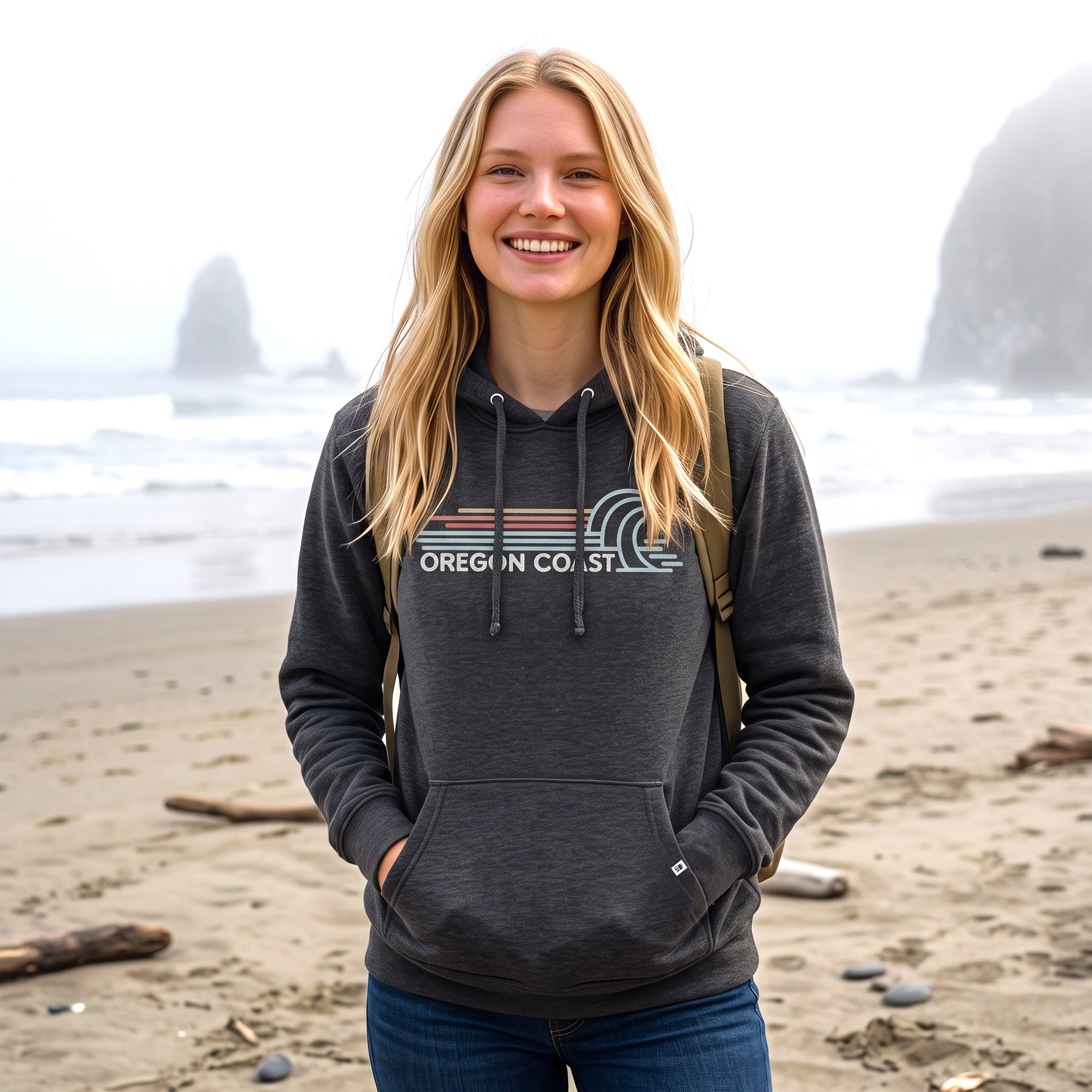 A smiling blonde woman stands on a sandy beach wearing a dark charcoal gray hoodie with a 'OREGON COAST' graphic across the chest, blue jeans, and a backpack.