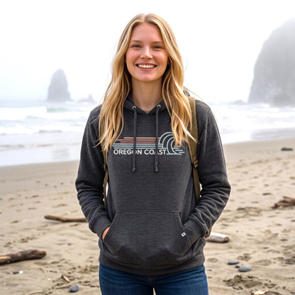 A smiling blonde woman stands on a sandy beach wearing a dark charcoal gray hoodie with a 'OREGON COAST' graphic across the chest, blue jeans, and a backpack.