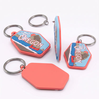 Four views of a silicone keychain featuring an Oregon border design, including a front view with a metal key ring, a side view, a back view with a blank coral-colored silicone panel, and another front view. The keychain has a blue and white Oregon border graphic with a mountain illustration, surrounded by a coral-red border, on a white background.