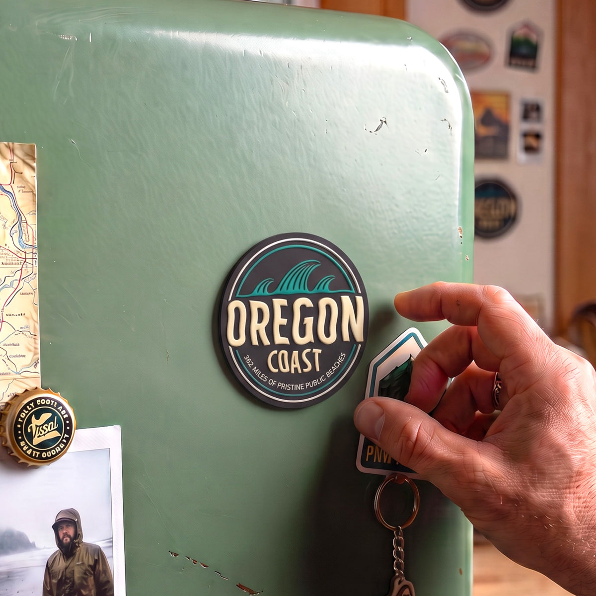 A hand is attaching a small Oregon Coast keychain to a mint green refrigerator. The keychain features a black circular magnet with teal and cream wave graphics and the text 'OREGON COAST', along with a small blue and green magnet on a metal key ring.
