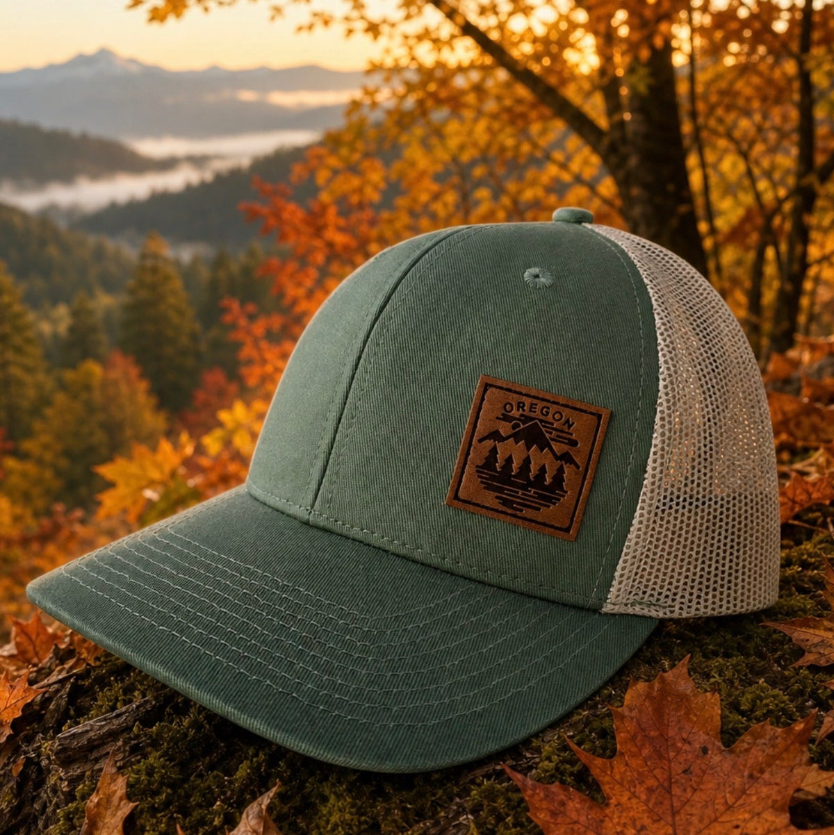 A green trucker-style cap with a brown leather patch logo, resting on a mossy log covered in autumn leaves, with a scenic mountain forest background at sunset.