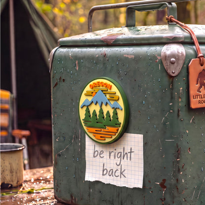 A round Oregon mountain-themed sticker or magnet is shown attached to a weathered green metal cooler outdoors, with a handwritten note reading 'be right back' below it.