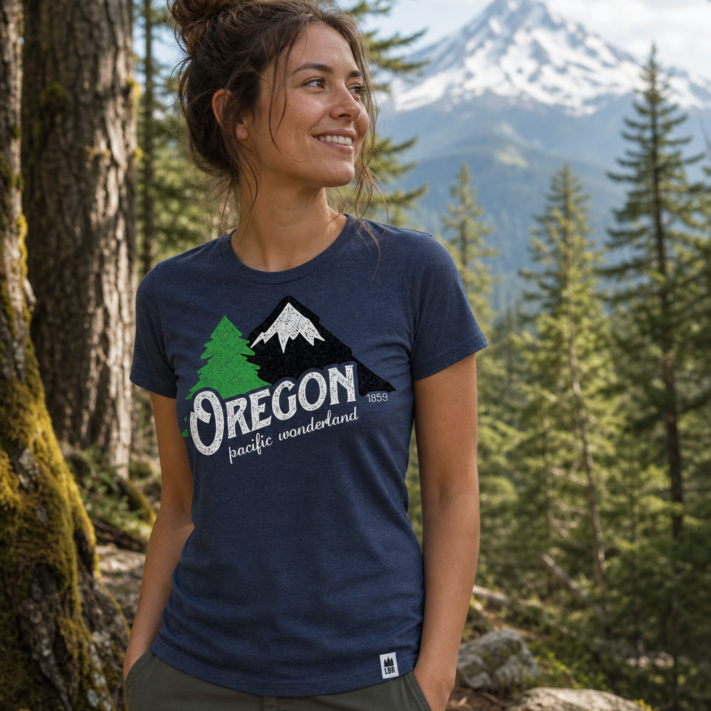 A woman wearing a navy blue graphic t-shirt with a green and white Oregon pine forest and mountain design, standing in a forest with mountains in the background.