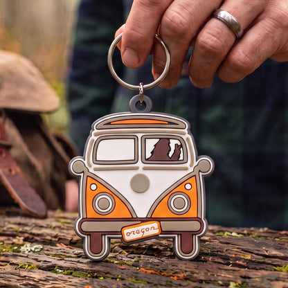 A hand holds a rubber keychain shaped like an orange and white vintage VW camper van with a metal key ring. The keychain has a small 'Oregon' sign on the front and is photographed outdoors on a log with a blurred rustic background.