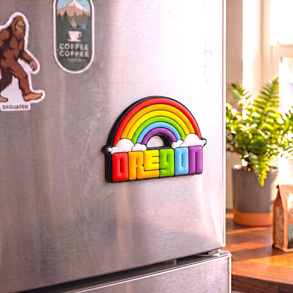 A colorful 3D rainbow refrigerator magnet spelling 'OREGON' with white clouds, attached to a stainless steel refrigerator in a kitchen setting.