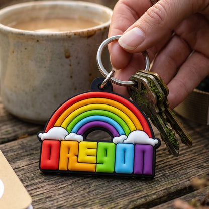 A hand holds a rainbow-shaped keychain with the word "OREGON" in colorful block letters, attached to metal keys, with a ceramic mug and wooden table in the background.