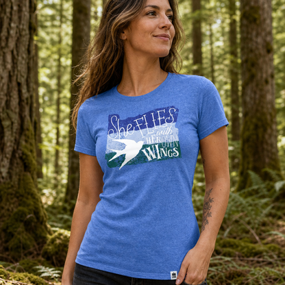 A woman wearing a heather blue graphic t-shirt with a bird design and the text “She Flies with her own Wings”' in a forest setting.