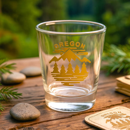 A clear glass shot tumbler with a gold Oregon mountain and tree design, set on a rustic wooden surface with green foliage in the background.