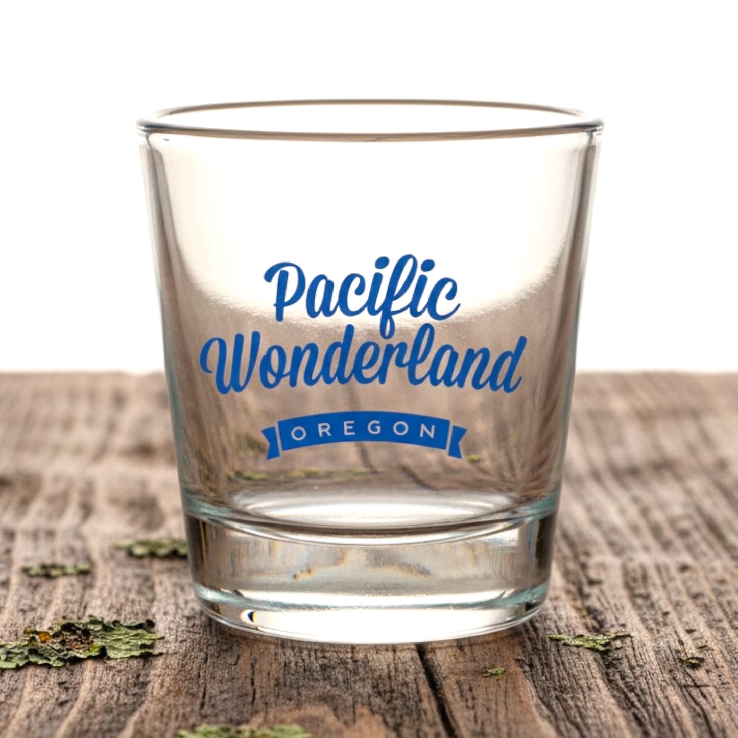 A clear glass tumbler with blue lettering that reads 'Pacific Wonderland OREGON,' shown on a rustic wooden surface against a white background.