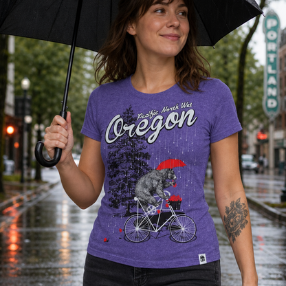 A woman wearing a purple graphic t-shirt with a raccoon on a bicycle under the text 'Pacific Northwest Oregon' and holding a black umbrella on a rainy city street.