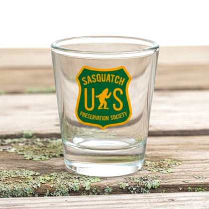 A clear glass shot glass featuring a green and yellow shield logo that reads 'SASQUATCH PRESERVATION SOCIETY’